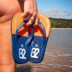 Person holding blue and orange flip-flops with a beach and water background