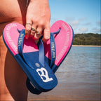 Person holding pink and blue flip-flops with a beach and water background