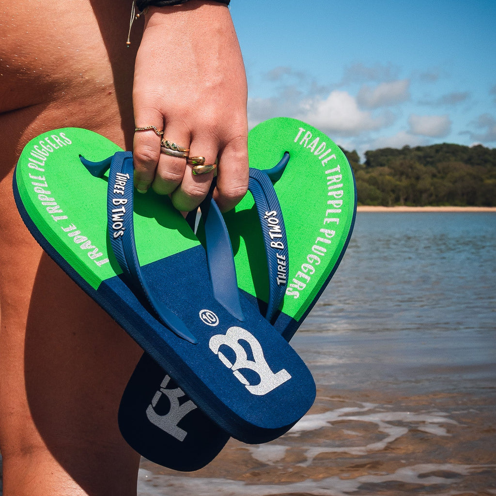 Person holding green and blue flip-flops by a body of water with a scenic background