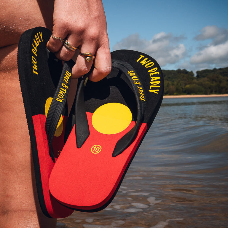 Person holding red and black sandals with yellow accents by a beach